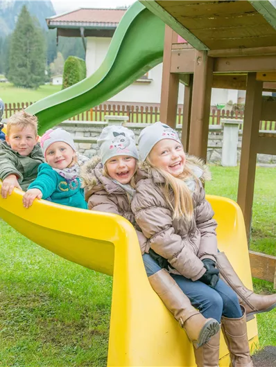Fünf Kinder spielen fröhlich auf einer Rutsche in einem schönen Spielplatz. Im Hintergrund sind grüne Wiesen und Bäume sichtbar.