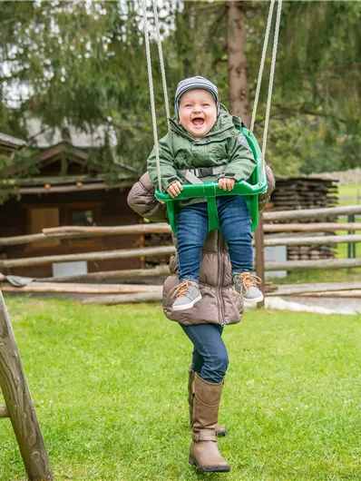 Ein fröhliches Kind schwingt auf einer Schaukel in einem grünen Garten. Im Hintergrund sind Bäume und Spielgeräte zu sehen.