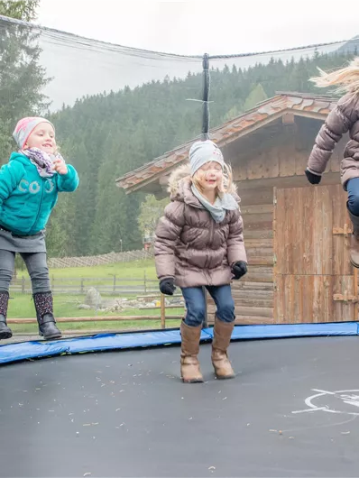 Drei Kinder springen fröhlich auf einem Trampolin. Im Hintergrund sind Bäume und ein Holzhaus zu sehen.
