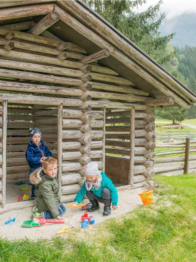Drei Kinder spielen vor einer Holzhütte auf einer Wiese. Im Hintergrund sind Bäume und Hügel sichtbar.