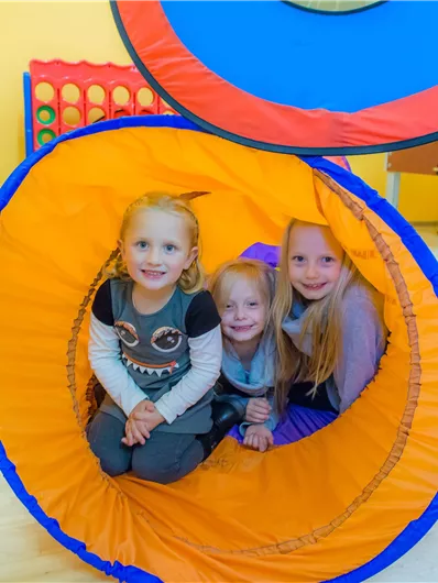 Drei fröhliche Kinder spielen in einem orangefarbenen Tunnel. Die Wände des Raumes sind gelb, und im Hintergrund sind mehrere Spielzeuge sichtbar.