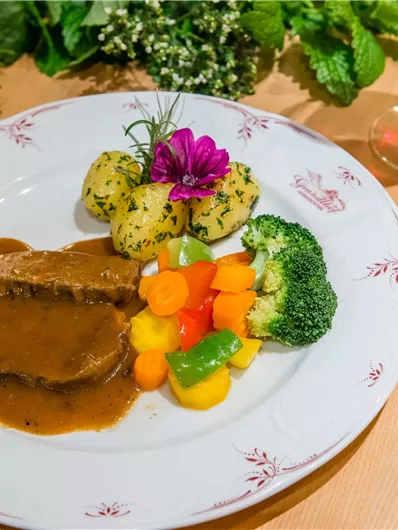 Ein leckeres Gericht mit zartem Fleisch in Bratensauce, Beilagen aus Gemüse und Kartoffeln. Daneben ein Glas Wasser und etwas Wein.