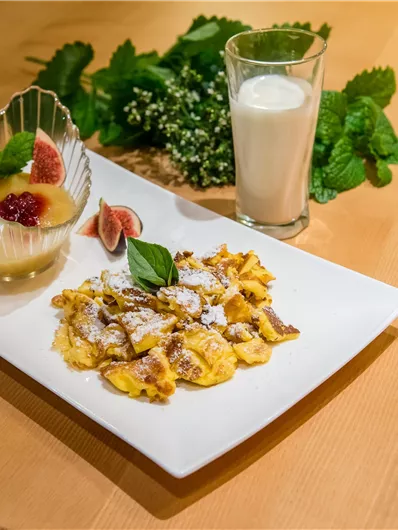 Ein Teller mit Kaiserschmarrn, garniert mit Puderzucker und frischen Früchten. Daneben steht ein Glas Milch und eine Dessertschale mit Obst.