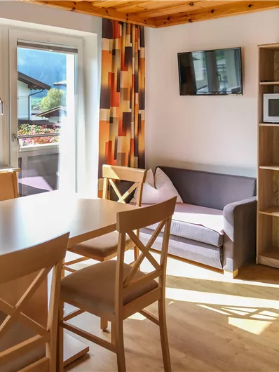 A bright and inviting holiday apartment with a dining area, a sofa, and a television. Large windows provide a view outside and ensure plenty of daylight.