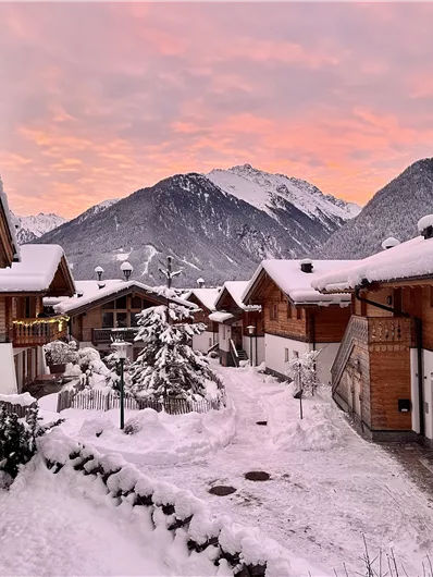 Eine winterliche Landschaft mit verschneiten Hütten und Berge im Hintergrund. Der Himmel erstrahlt in warmen Farbnuancen beim Sonnenuntergang.