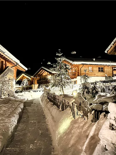 Ein malerisches Dorf im Schnee bei Nacht, umgeben von hölzernen Häusern. Der Weg ist von frischem Schnee bedeckt und sorgt für eine gemütliche Winteratmosphäre.