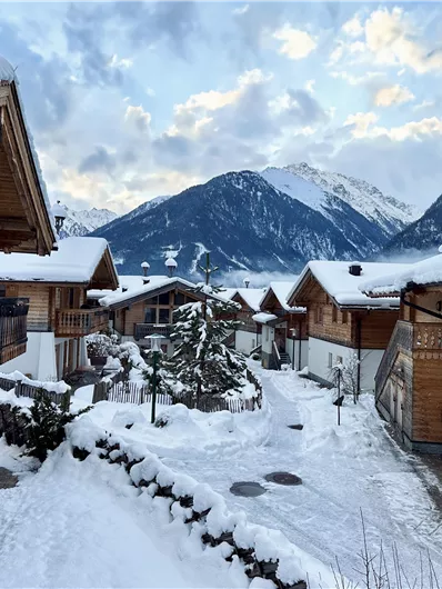 Ein malerisches Winterdorf mit Holzhäusern und schneebedeckten Wegen. Im Hintergrund erheben sich majestätische Berge unter einem klaren Himmel.