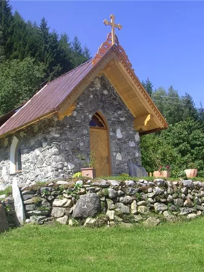 Eine kleine Steinkapelle umgeben von Grünflächen und Bäumen. Im Hintergrund sind die Berge sichtbar und der Himmel ist blau.