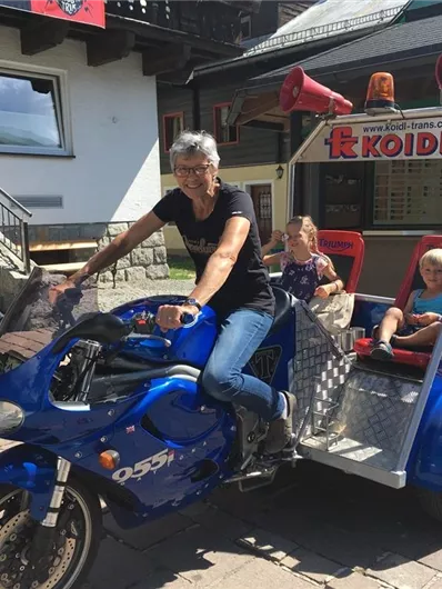 Een vrouw zit op een blauwe motorfiets met een aanhangwagen, waarin twee kinderen zitten. Op de achtergrond zijn een trap en een gebouw te zien.