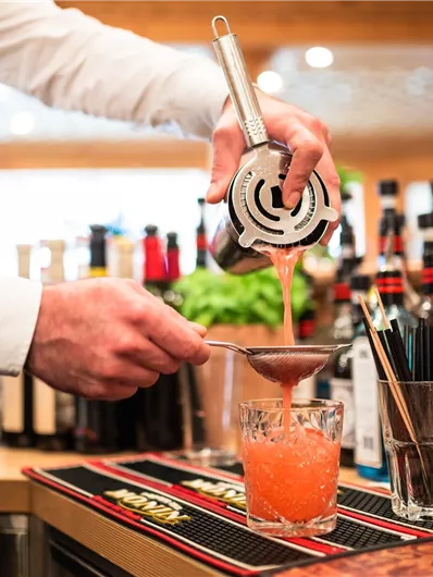 Ein Barkeeper gießt einen bunten Cocktail in ein Glas. Im Hintergrund sind verschiedene Flaschen und Utensilien sichtbar.