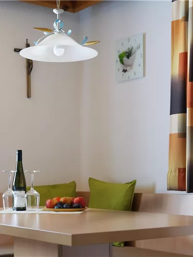 A cozy dining area with a wooden table and two chairs. On the table are wine and fruit, and the windows are decorated with colorful curtains.
