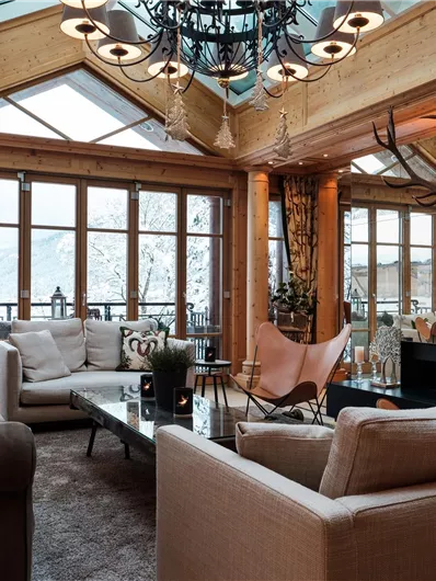 Ein stilvolles Wohnzimmer mit großen Fenstern und einem Blick auf die schneebedeckten Berge. Die Einrichtung ist gemütlich und aus Holz, mit modernen Möbeln und dekorativen Elementen.