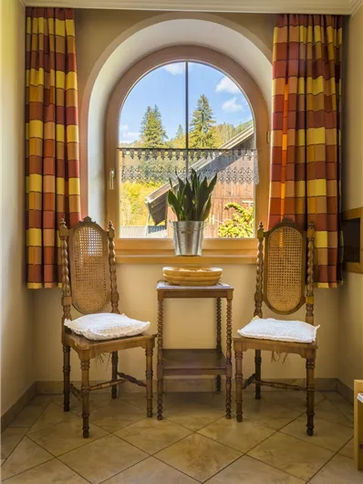 Een lichte kamer met twee stoelen en een kleine tafel. Bij het raam staan planten en er zijn decoratieve gordijnen.