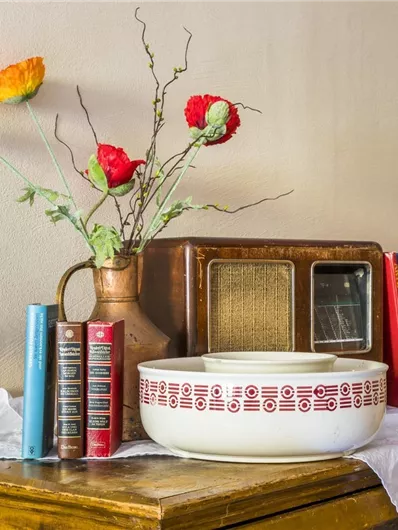 Een houten kast met een vintage radio, een porseleinen kom en kleurige bloemen in een vaas. Naast de radio staan verschillende boeken in diverse kleuren.