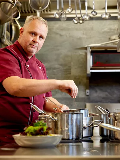 Een kok in een moderne keuken bereidt een gerecht. Hij staat voor een fornuis met potten en een kom verse groenten.