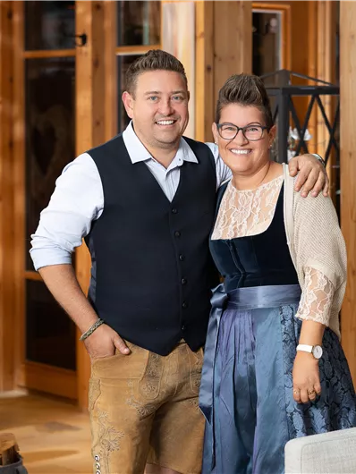 Ein Paar steht lächelnd in einem gemütlichen, hölzernen Raum. Der Mann trägt eine Weste und die Frau ein elegantes Kleid.