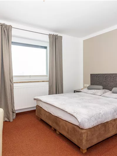 Een eenvoudige slaapkamer met een tweepersoonsbed en een stoel. Grote ramen laten veel licht binnen.