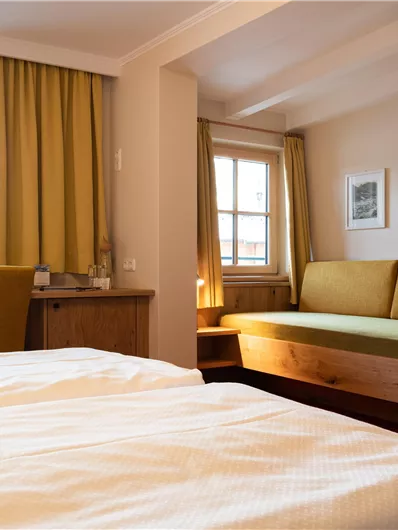 Ein gemütliches Zimmer mit einem Doppelbett, einem Sofa und einem Schreibtisch. Große Fenster sorgen für viel Licht und eine angenehme Atmosphäre.