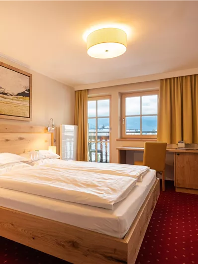 Ein gemütliches Hotelzimmer mit einem Doppelbett und Holzmöbeln. Große Fenster sorgen für viel Tageslicht und Blick nach draußen.