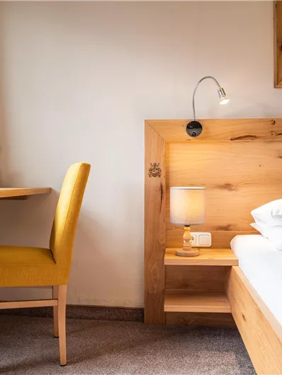 Ein gemütliches Zimmer mit einem Holzbett und einem einfachen Schreibtisch. Gelbe Vorhänge lassen viel Tageslicht herein.