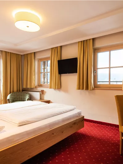 Ein gemütliches Hotelzimmer mit einem Doppelbett und hellen Vorhängen. Der Raum ist mit einem Schreibtisch, einem Fernseher und großen Fenstern ausgestattet.