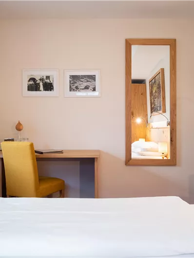 Ein gemütliches Zimmer mit einem Bett, einem Schreibtisch und einem Stuhl. Der Raum hat helle Wände und ein großes Fenster mit Vorhängen.