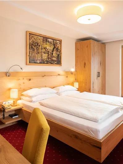 Ein gemütliches Hotelzimmer mit einem Doppelbett und einem Tisch. Das Zimmer hat helle Möbel und große Fenster mit Blick nach draußen.