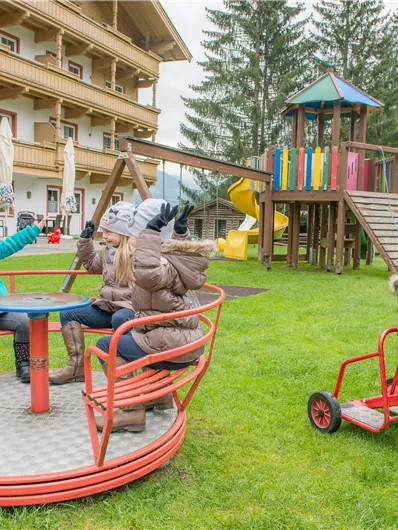 Ein Spielplatz mit Kindern, die auf einem Karussell sitzen und spielen. Im Hintergrund sind ein Spielhaus und eine Rutsche zu sehen.