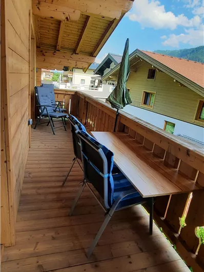 Een gezellige balkon met twee stoelen en een tafel. Het landschap is omgeven door houten huizen en een heldere, blauwe lucht.