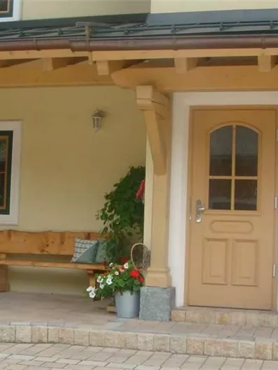 Een uitnodigende ingang van een huis met een houten bank en bloeiende planten. De muren zijn geschilderd in een warme gele tint en de deuren zijn in een vriendelijke beige kleur.