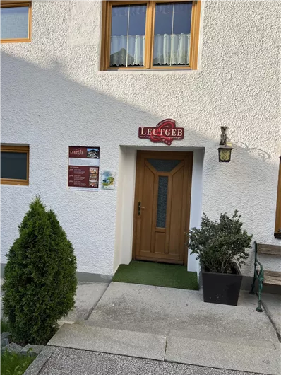 An inviting entrance to a building featuring a wooden door and a sign that says "Leutgeb." On the side, there are plants, and a light fixture is mounted above the door.