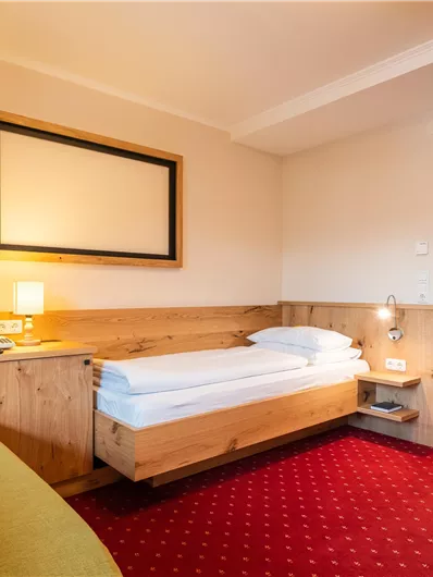 Ein einfaches Hotelzimmer mit einem Einzelbett, einem Nachttisch und einem großen Spiegel an der Wand. Der Boden ist rot, und die Wände sind hell gehalten.
