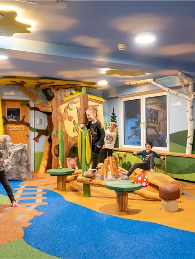 Ein bunter Spielraum mit Spielgeräten, wie einem Baumhaus und mehreren Tieren. Kinder spielen fröhlich auf dem weichen Bodenbelag.
