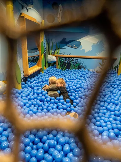 Ein kleines Kind spielt in einem Pool aus blauen Bällen. Im Hintergrund sind Fische und Pflanzen in einer bunten Wandmalerei zu sehen.