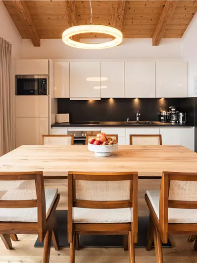 Eine moderne Küche mit einem großen Holztisch und Stühlen. Tageslicht strömt durch die Fenster und sorgt für eine gemütliche Atmosphäre.