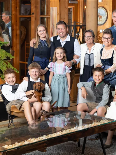 Eine fröhliche Familie posiert in einem gemütlichen Raum. Sie stehen und sitzen um einen Tisch mit einer Holzoberfläche, umgeben von stilvollen Möbeln.