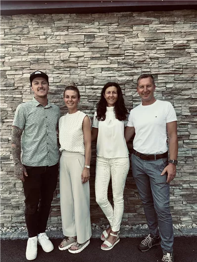 A group of four people is standing in front of a stone wall. They are smiling and wearing trendy, light-colored clothing.