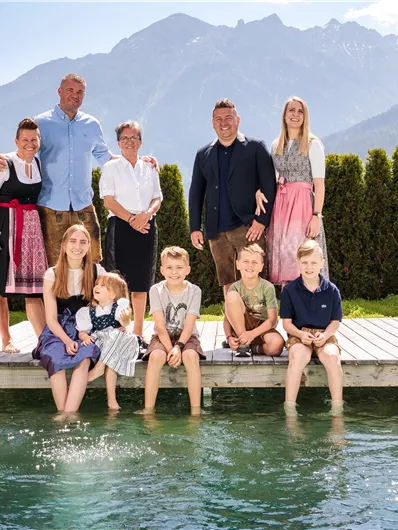 Eine fröhliche Gruppe von neun Personen steht auf einem Steg am Wasser mit Bergen im Hintergrund. Alle Teilnehmer tragen traditionelle Trachten, und es herrscht eine freundliche, festliche Atmosphäre.