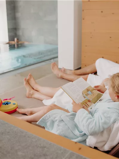Eine entspannte Familie sitzt am Beckenrand und genießt die Zeit miteinander. Ein Kind liest ein Buch, während die Erwachsenen sich zurücklehnen und entspannen.