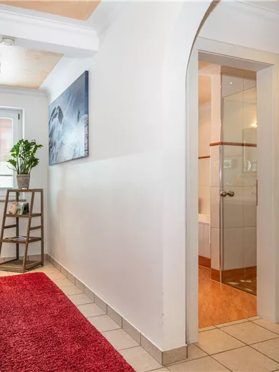 Een heldere gang met een rood tapijt en een plantenstandaard. Op de achtergrond is een badkamer zichtbaar.