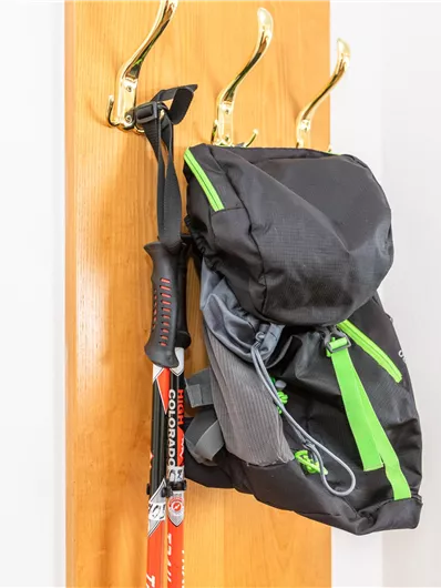 Een rugzak hangt aan een kapstok naast een wandelstok. De rugzak is zwart met groene accenten.