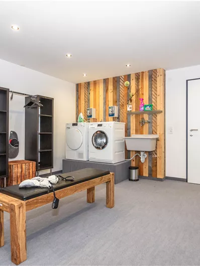 Een moderne wasruimte met een wasmachine, een wastafel en houten planken. De inrichting is functioneel en uitnodigend vormgegeven.