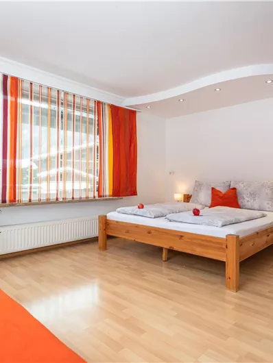 Een heldere slaapkamer met een groot tweepersoonsbed en oranje gordijnen. Er is een televisie en een kleine plant in de kamer.