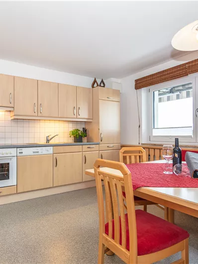 Een moderne keuken met houten kasten en lichte muren. Een eettafel met stoelen en een raam dat veel daglicht binnenlaat.