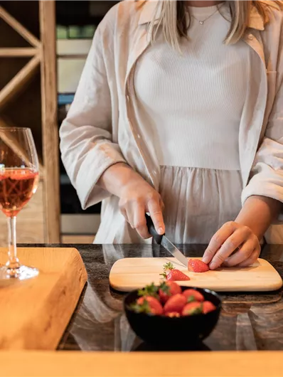 A person is cutting fresh tomatoes on a wooden table in the kitchen. In the background, there is a glass of rosé wine and a bowl of strawberries.