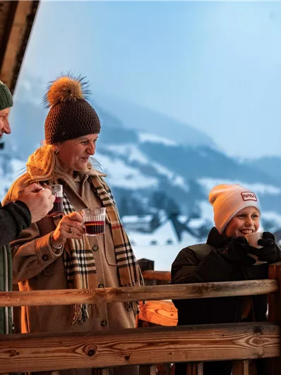 Three people are standing on a balcony, enjoying the view of a winter landscape. They are warmly dressed and holding drinks in their hands.