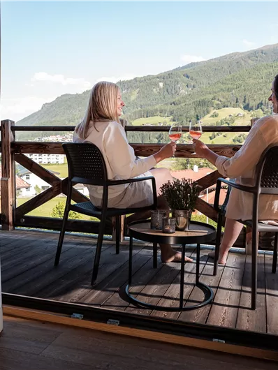 Two women are sitting on a balcony and enjoying the view of the mountains. They are wearing bathrobes and relaxing in the peaceful atmosphere.