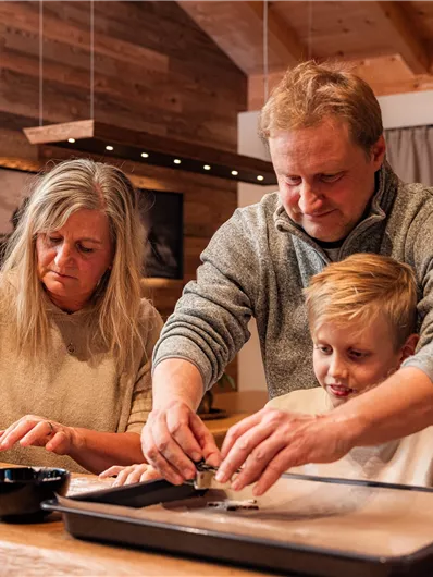 A family is preparing food together in a cozy kitchen environment. All three are focused on preparing ingredients.