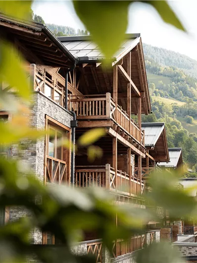 A wooden building with balconies in a mountainous landscape. In the foreground, green leaves are visible, forming a natural frame.