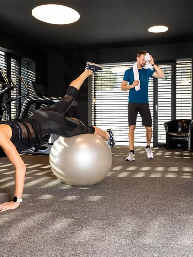 A woman is training on a gym ball while a man in sportswear cheers her on. The room has modern fitness equipment and plenty of daylight.